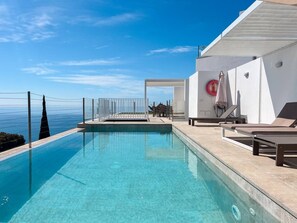 Une piscine d’un bleu éclatant, avec vue sur la mer, un bâtiment blanc orné d’une bouée de sauvetage et un parasol.