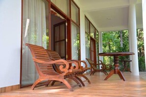 A covered outdoor seating area with wooden benches and a round table, surrounded by large windows. - River Edge (Thissamaharama)