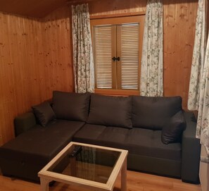 A wooden-paneled room with a sectional sofa, a glass coffee table, and a window with shutters.