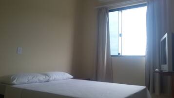 A single-bed room with a bed, a window with curtains, an air conditioning unit, and a television on a stand.
