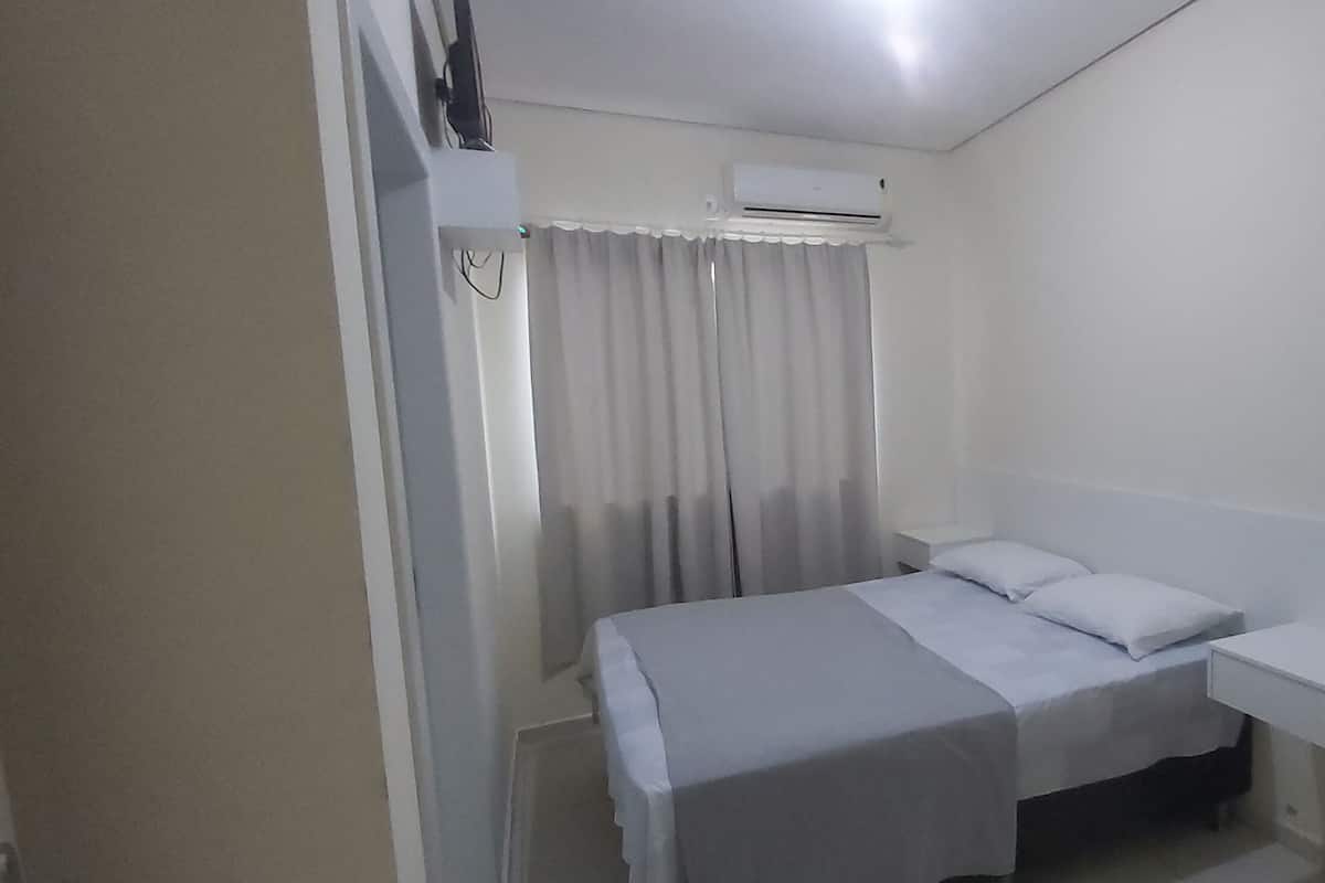 A small, clean room with a bed, a bedside table, a chair, and an air conditioning unit.