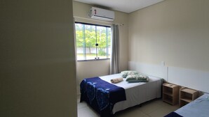 A small, single-bed room with a window, a bedside table, and a wall-mounted air conditioner. - Pousada Chef Brasil (Barra Velha)