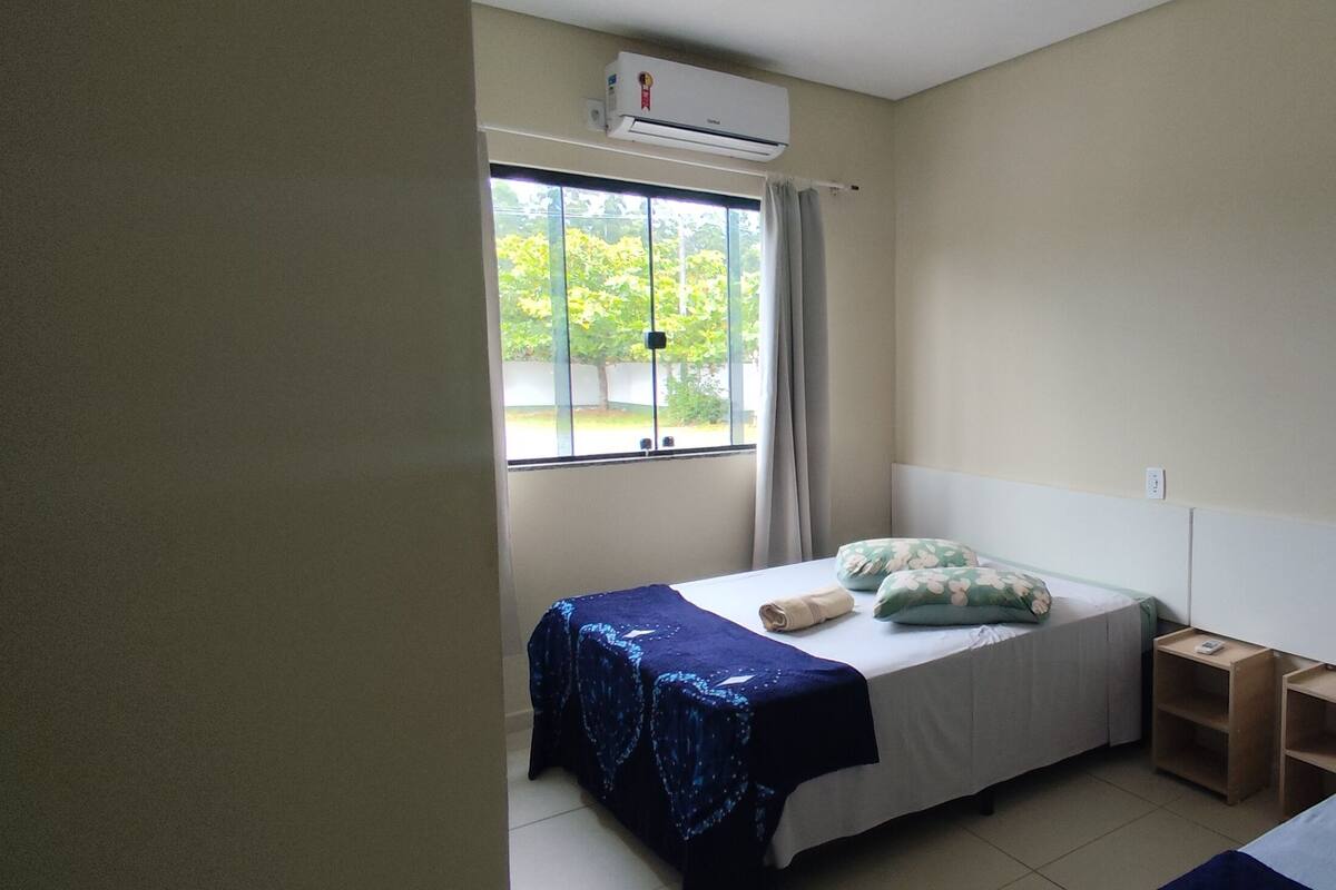 A small, single-bed room with a window, a bedside table, and a wall-mounted air conditioner.