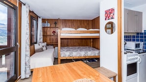 Quarto compacto com beliche, pequeno espaço de cozinha, mesa de madeira e vista por uma grande janela.