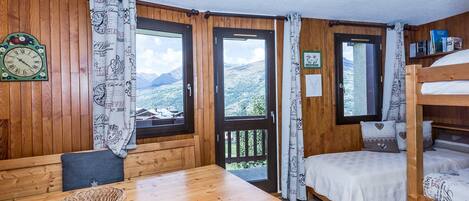 A wooden cabin with a dining table, bunk beds, and large windows offering mountain views.