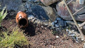 Eine Steinsäule mit einer Holzleiter, umgeben von Grünpflanzen und Gartenmöbeln.
