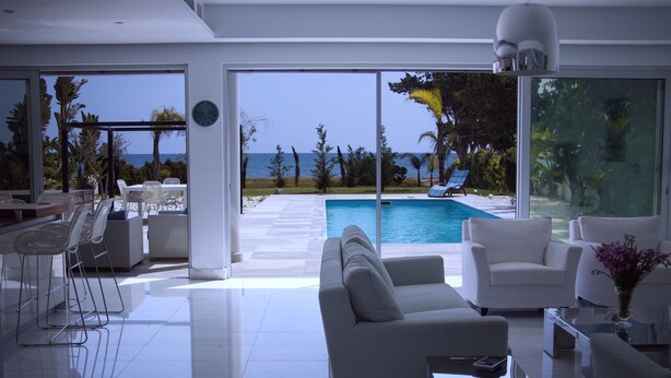 Un soggiorno moderno con un'ampia porta a vetri che conduce a una zona piscina, arredo bianco e vista sull'oceano.