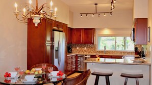 A modern kitchen with wooden cabinets, a central island with bar stools, a chandelier, and a dining area with a table set for two. - Beautiful Vista Waikoloa B105 - Renovated in 2018! (Waikoloa)