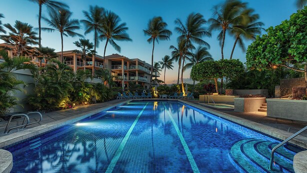 A swimming pool with clear blue tiles, surrounded by palm trees and a building with balconies. - Beautiful Vista Waikoloa B105 - Renovated in 2018! (Waikoloa)
