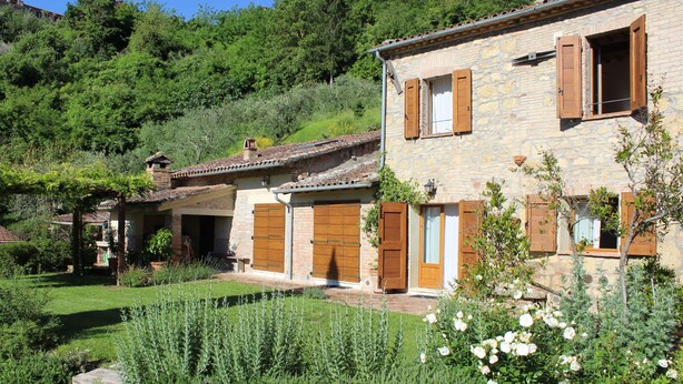 Una casa in pietra con persiane di legno, un giardino con rose e una collina sullo sfondo.