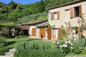 Una casa de piedra con contraventanas de madera, un jardín de rosas y una colina al fondo.
