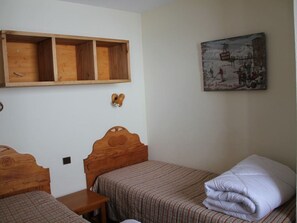 Quarto com duas camas de madeira, uma prateleira de madeira acima das camas e um candeeiro fixado na parede.