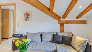 Une pièce avec un plafond en bois, un canapé gris avec des coussins, une petite table sur laquelle est posé un vase de fleurs, et une porte qui donne sur une autre pièce.