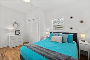 Quarto com cama, mesas de cabeceira, cômoda, porta e um objeto decorativo na parede.