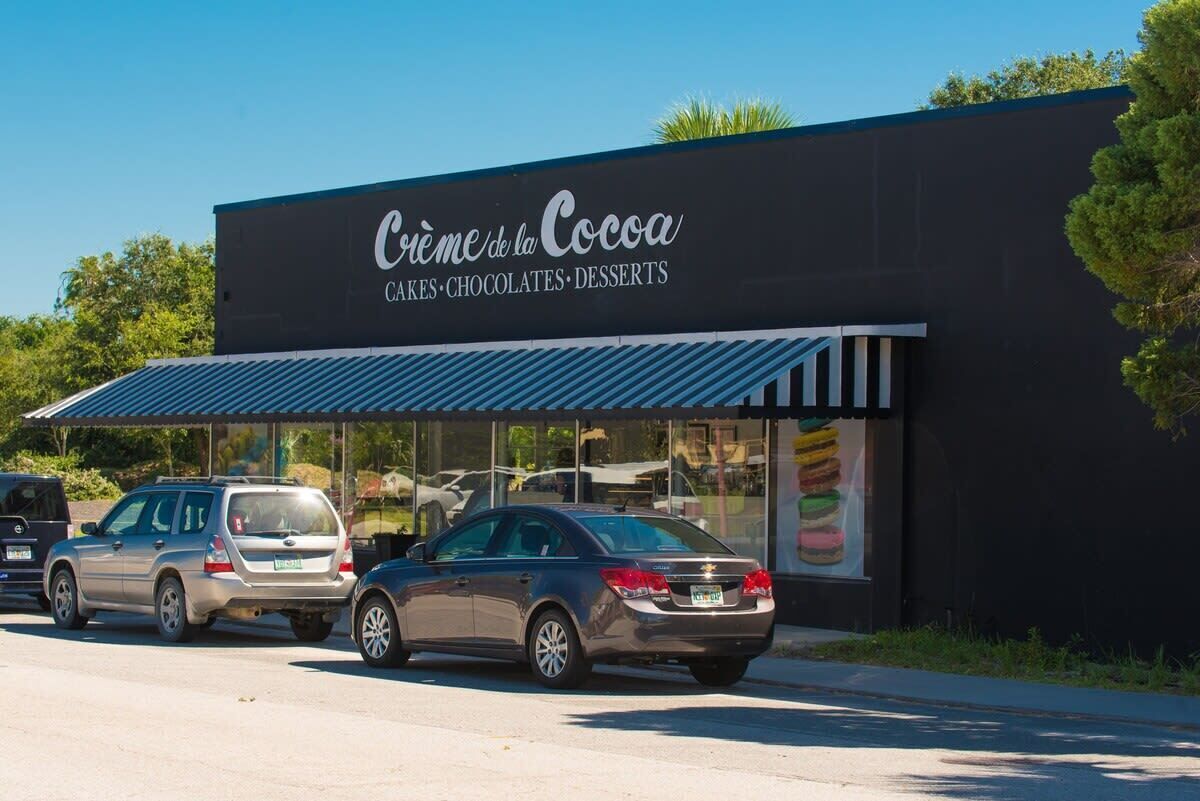 A storefront with a sign reading 'Crème de la Cocoa' and a menu of cakes, chocolates, and desserts.