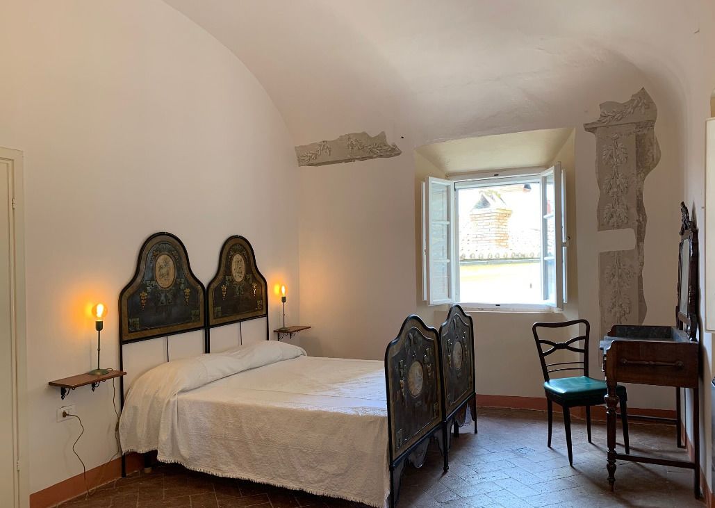 A bedroom with a bed, two wall-mounted lamps, a chair, a desk, and a window with a view of the outside.
