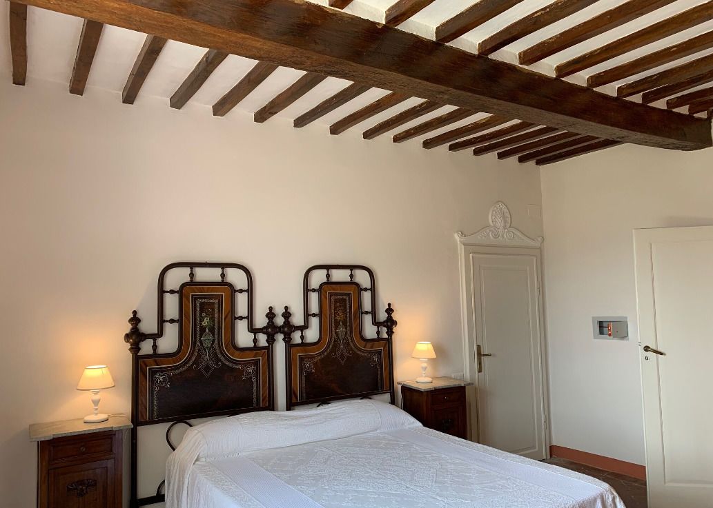 A bedroom with a wooden ceiling, two ornate headboards, a white bedspread, two bedside lamps, a nightstand, and a door.