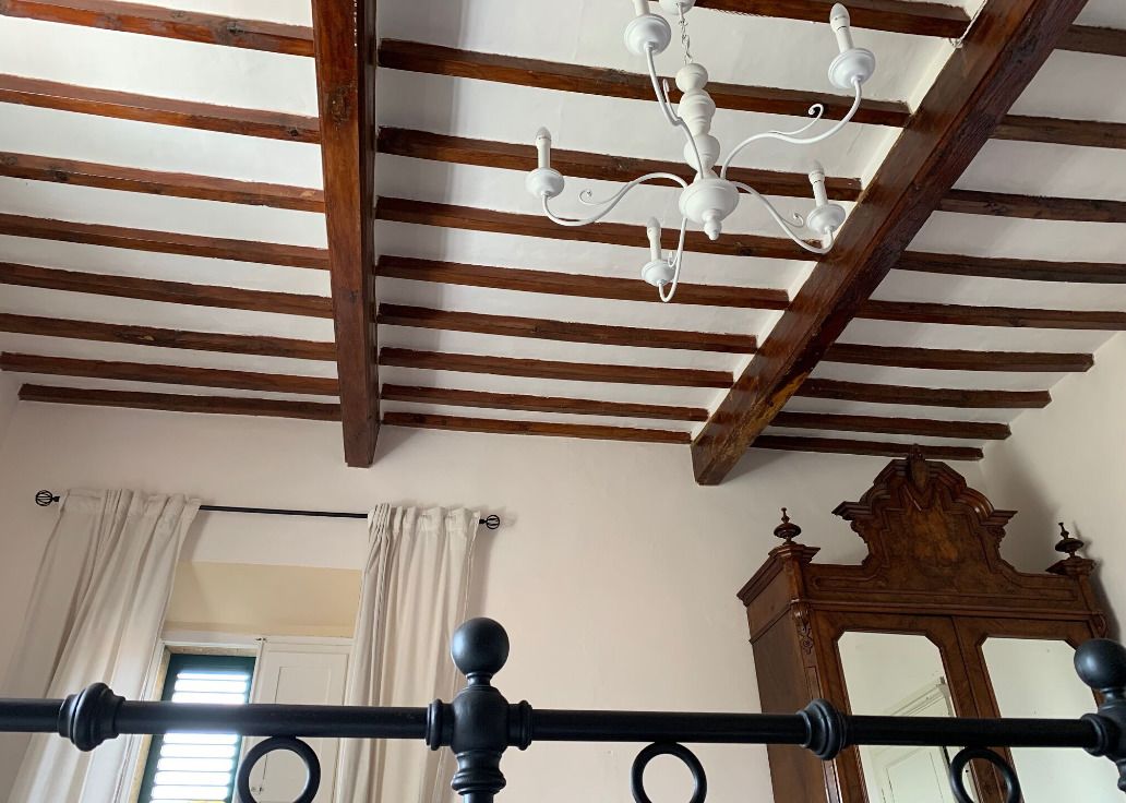 Ein Zimmer mit Holzbalkendecke, einem Chandelier, einem Bett mit schwarzem Kopfteil und einem Holzkabinett.