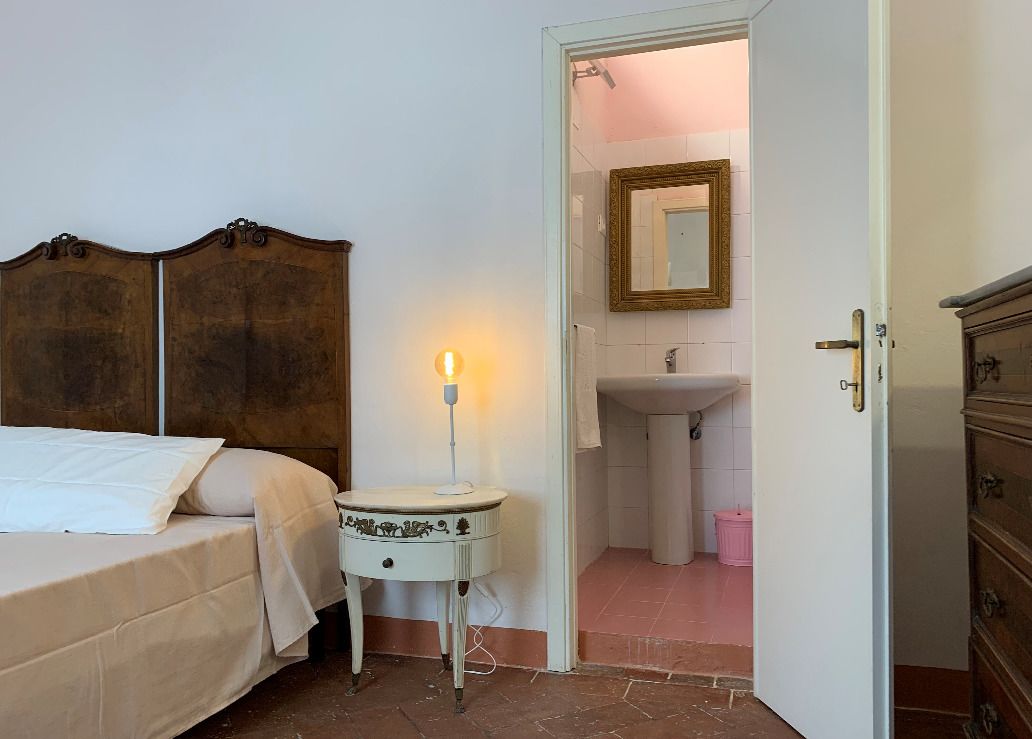 A bedroom with a bed, bedside table with a lamp, a wooden headboard, and a door leading to a bathroom with a sink and mirror.