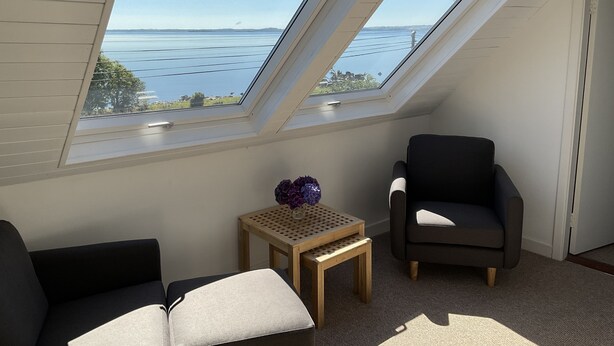 A room with a sloped ceiling and skylights, a dark armchair, a small wooden table with a vase of flowers, and a view of a body of water and distant land. -  Lakeside Lough Mask shore (Tourmakeady)