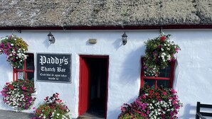 A white building with a red door and windows, adorned with hanging flower baskets. A sign above the door reads "Paddy's Thatch Bar." -  Lakeside Lough Mask shore (Tourmakeady)