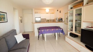 A compact living space with a sofa, a dining table with a purple tablecloth, a kitchen area with cabinets and a refrigerator, and a television on a stand. - Comfortable apartment near the beach and services (Saint-Raphaël)