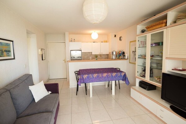 A compact living space with a sofa, a dining table with a purple tablecloth, a kitchen area with cabinets and a refrigerator, and a television on a stand.