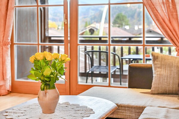 A room with a large window, a vase with yellow flowers, a doily, and a sofa with a cushion.