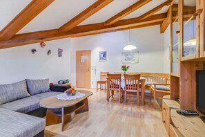 A cozy living room with a sofa, a wooden dining table with chairs, a television, and a ceiling with wooden beams.