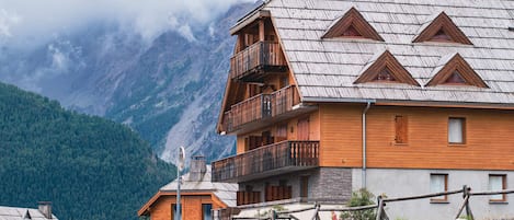 A wooden chalet with a balcony, set against a mountainous backdrop.