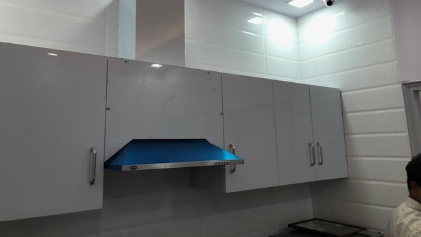 A modern kitchen with white cabinets, a blue exhaust hood, and recessed ceiling lighting. - Atul's Europa Apartments (New Delhi)