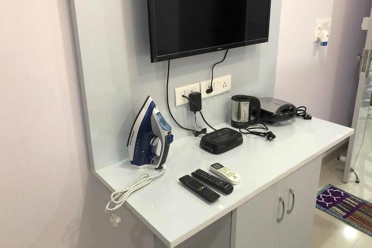 A white desk with a flat-screen TV, an iron, a kettle, a phone, and two remote controls.