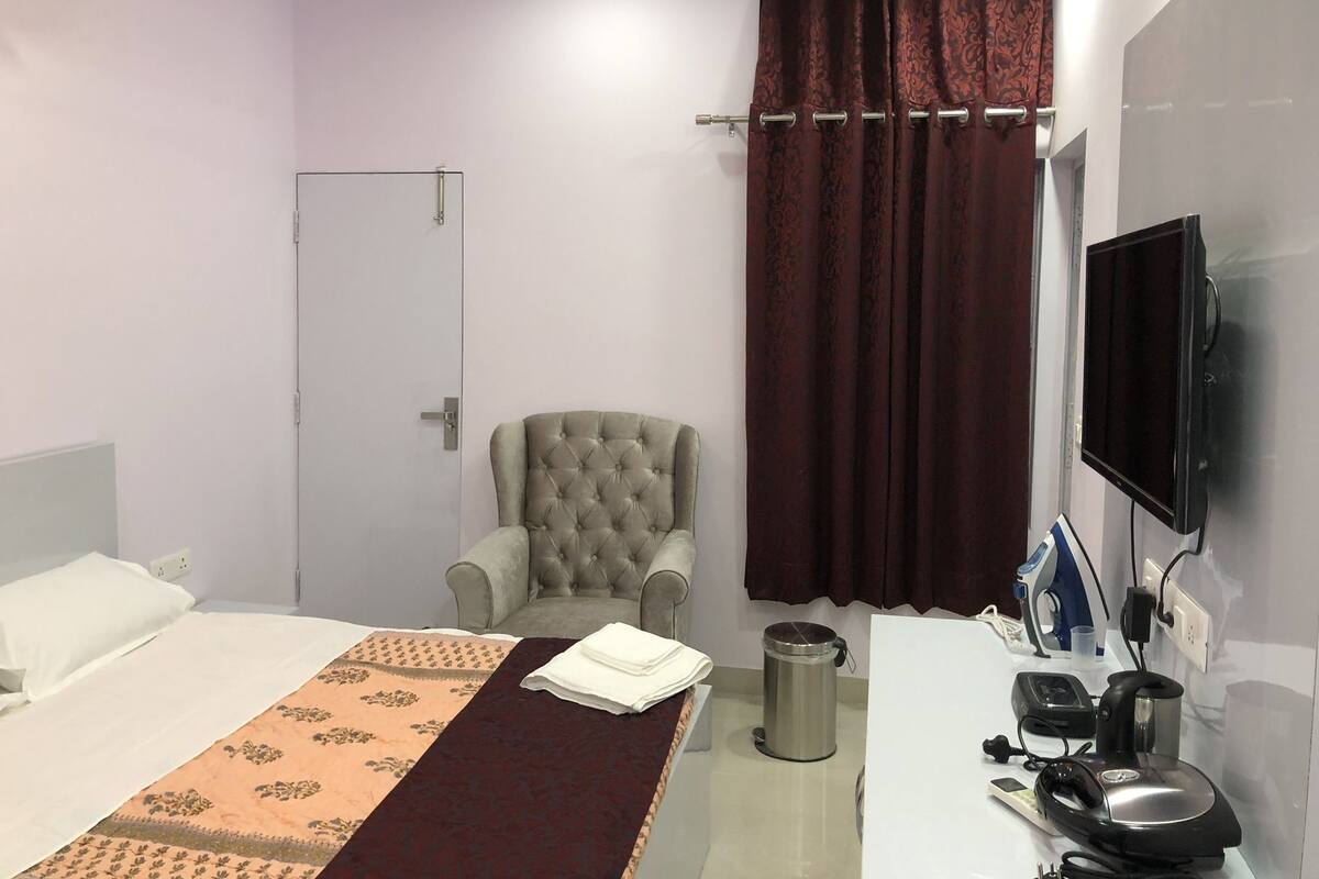 A hotel room with a bed, a grey armchair, a TV, a desk with a kettle, and a trash bin.
