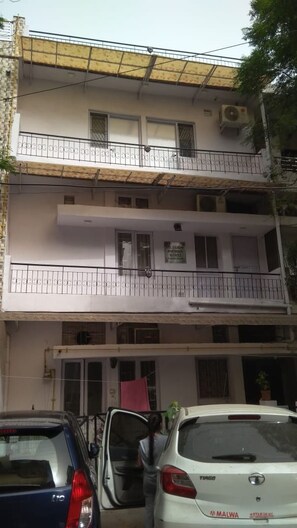 A multi-story building with balconies, a car parked in front, and a sign indicating a business on the ground floor.