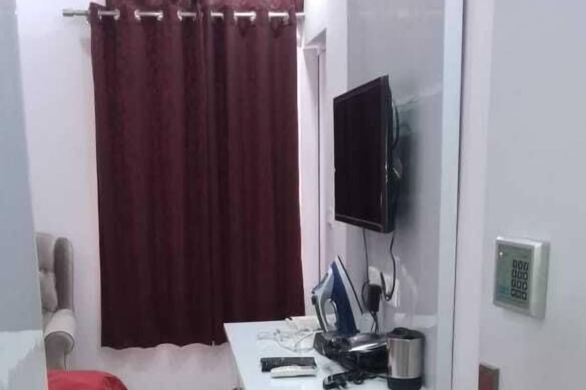 A small, compact room with a desk, a television mounted on the wall, a red curtain, and a trash bin.