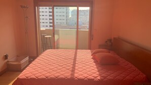 A bedroom with a large bed, a nightstand, a lamp, a small table, and a view of buildings through a sliding door. - Apartment Braga Parque (Braga)