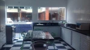 A kitchen with a checkered floor, a dining table with chairs, a refrigerator, and a window overlooking a brick building. - GATED COMMUNITY, 24-HOUR SECURITY, SWIMMING POOL WITH HYDROMASSAGE.  (Peruíbe - SP)