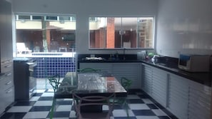 Uma cozinha com chão xadrez, mesa de jantar com cadeiras, geladeira e janela com vista para um prédio de tijolos.