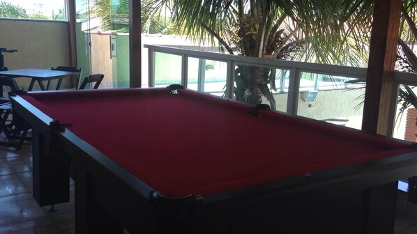 A pool table with a red felt surface, surrounded by chairs and tables, in a room with a glass railing and a view of palm trees outside. - GATED COMMUNITY, 24-HOUR SECURITY, SWIMMING POOL WITH HYDROMASSAGE.  (Peruíbe - SP)