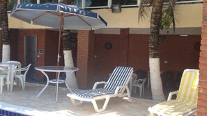 An outdoor seating area with a table and two lounge chairs, one striped and one yellow and white striped, under a blue umbrella. - GATED COMMUNITY, 24-HOUR SECURITY, SWIMMING POOL WITH HYDROMASSAGE.  (Peruíbe - SP)