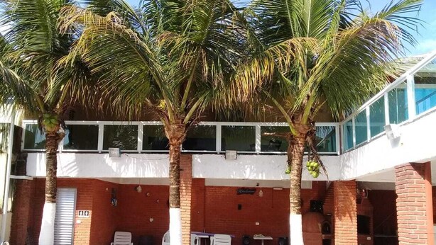 A swimming pool with palm trees, a brick building, and a covered area with chairs and speakers. - GATED COMMUNITY, 24-HOUR SECURITY, SWIMMING POOL WITH HYDROMASSAGE.  (Peruíbe - SP)