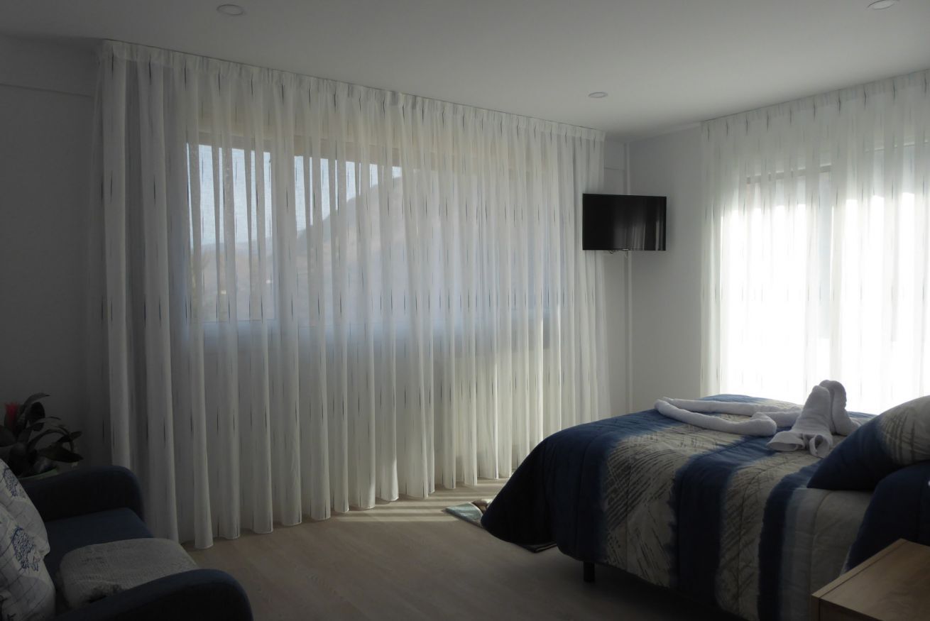 A hotel room with a bed, a TV mounted on the wall, and sheer curtains.