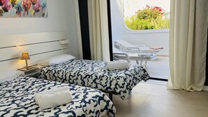 A hotel room with two beds, a nightstand, a lamp, and a view of the outside balcony with chairs. - Americas Holiday Home (Playa de las Américas)