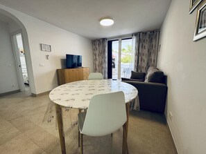 Une petite pièce bien éclairée, avec une table ronde, deux chaises, un canapé, un téléviseur et une porte-fenêtre coulissante donnant sur un espace extérieur.