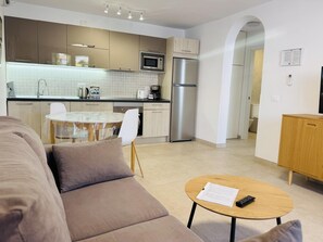 Ein modernes Wohnzimmer mit einer Couch, einem Couchtisch mit Fernbedienung und einer Küchenzeile mit Geräten sowie einem Esstisch.