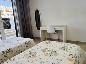 A room with two beds, a desk, a chair, a window with curtains, and a view of buildings outside. - Americas Holiday Home (Playa de las Américas)