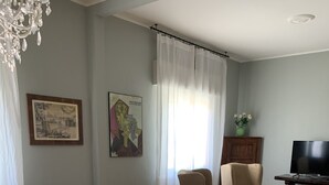 Uma sala de estar com piso de madeira em padrão herringbone, uma mesa redonda de madeira, uma poltrona bege, um sofá, uma televisão sobre suporte de madeira e um lustre.