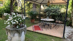 Alpendre coberto com mesa e cadeiras, rodeado por vegetação e um vaso de pedra decorativo com flores brancas.