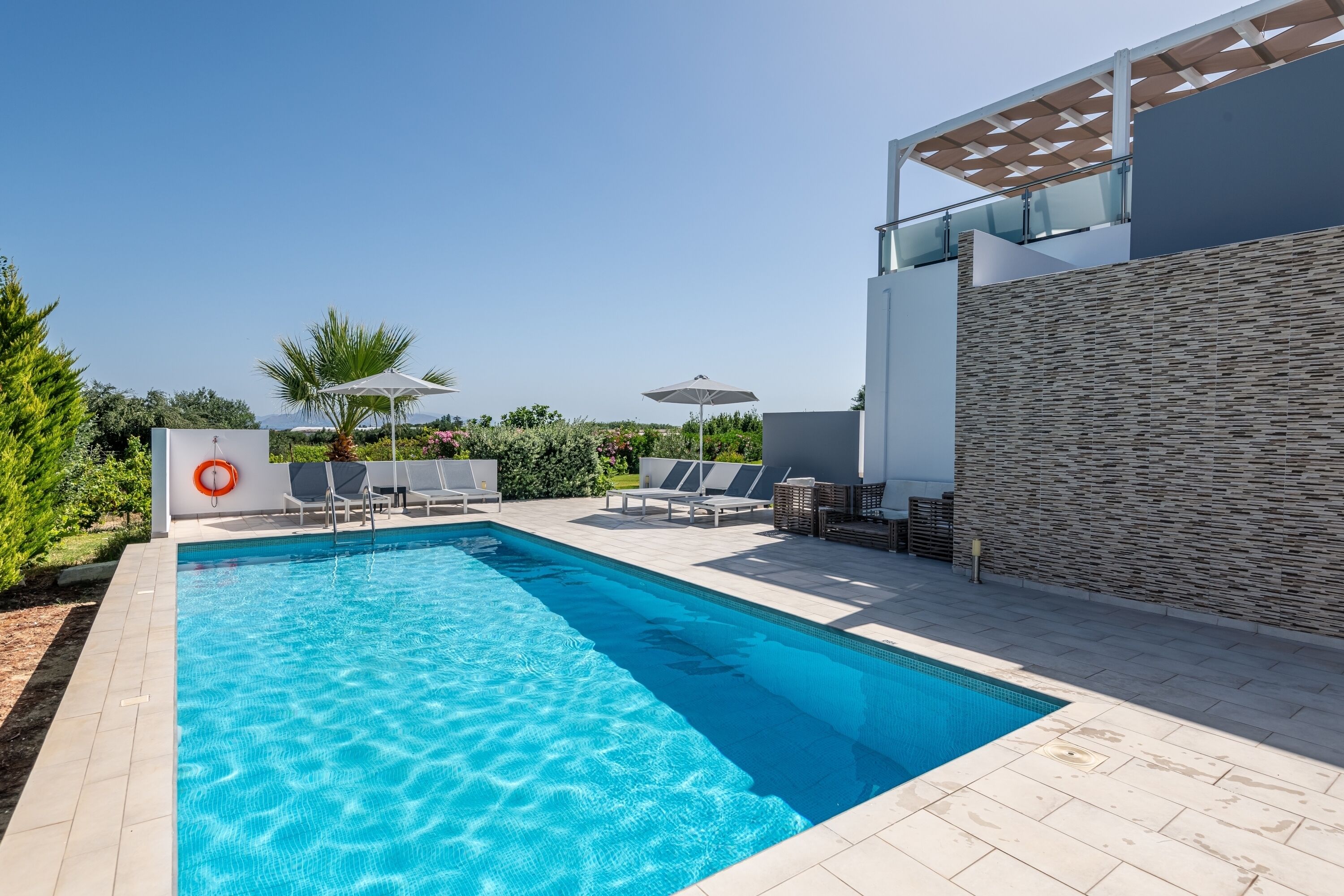 Ein Swimmingpool mit Liegestühlen und Sonnenschirmen, umgeben von einem modernen Gebäude mit einer Steinaußenwand.