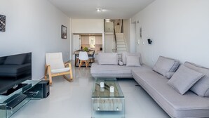 A modern living room with a sofa, a glass coffee table, a TV, and a dining area in the background.